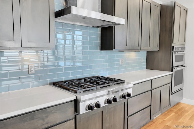 kitchen featuring appliances with stainless steel finishes, light wood-type flooring, backsplash, wall chimney range hood, and gray cabinets