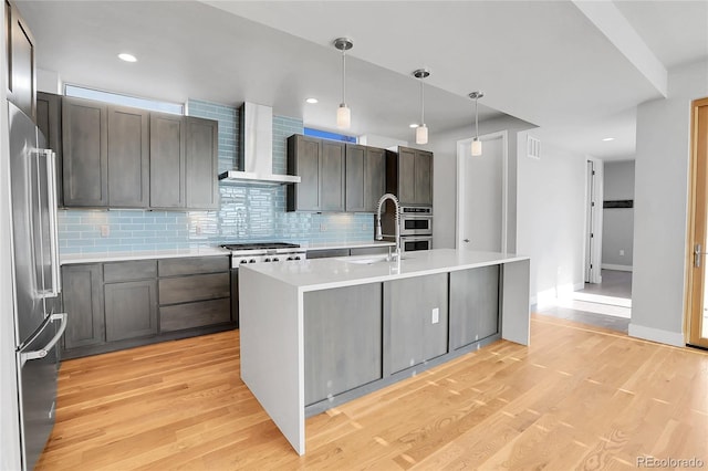 kitchen with pendant lighting, a center island with sink, wall chimney exhaust hood, light hardwood / wood-style floors, and stainless steel appliances