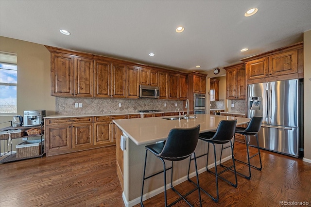 kitchen with a kitchen island with sink, stainless steel appliances, dark wood-style flooring, a sink, and light countertops