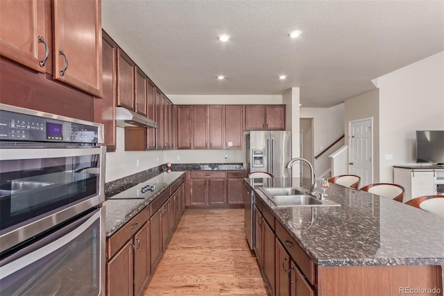 kitchen featuring a sink, stainless steel appliances, under cabinet range hood, light wood finished floors, and an island with sink