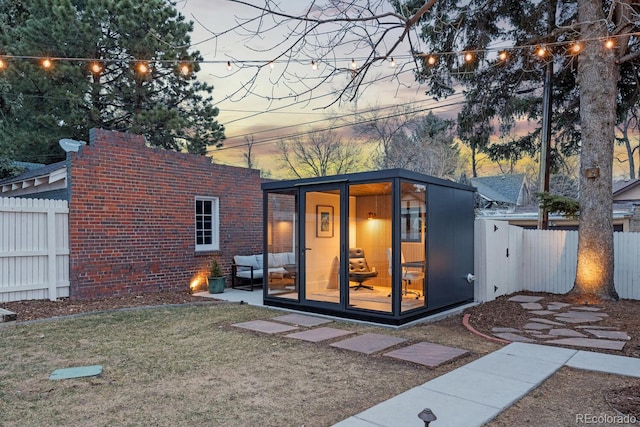 back of property at dusk featuring brick siding, fence, an outbuilding, and a yard