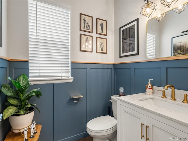 half bathroom with toilet, a wainscoted wall, vanity, and a decorative wall