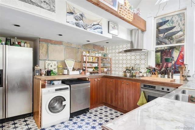 kitchen featuring stainless steel appliances, brown cabinetry, wall chimney range hood, light floors, and washer / dryer