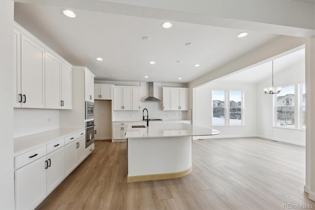 kitchen featuring a center island with sink, wall chimney exhaust hood, stainless steel appliances, light hardwood / wood-style floors, and white cabinetry