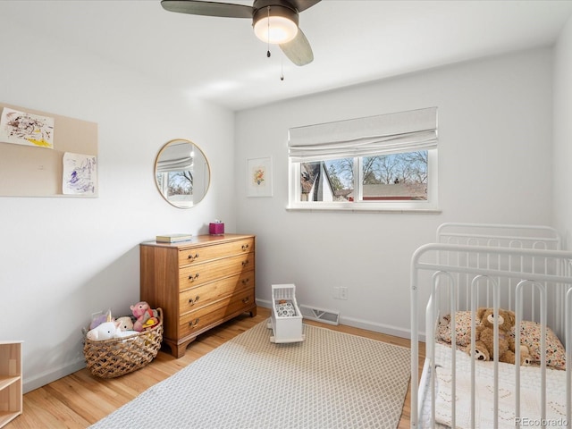bedroom featuring light wood-type flooring, baseboards, ceiling fan, a crib, and visible vents