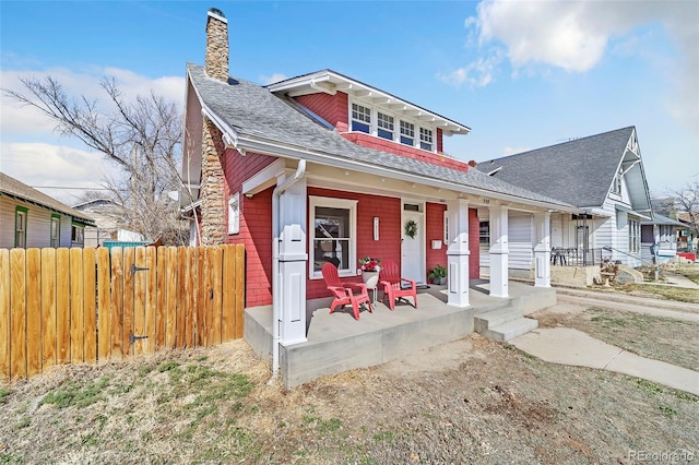 view of front facade featuring a porch, a shingled roof, a chimney, and fence