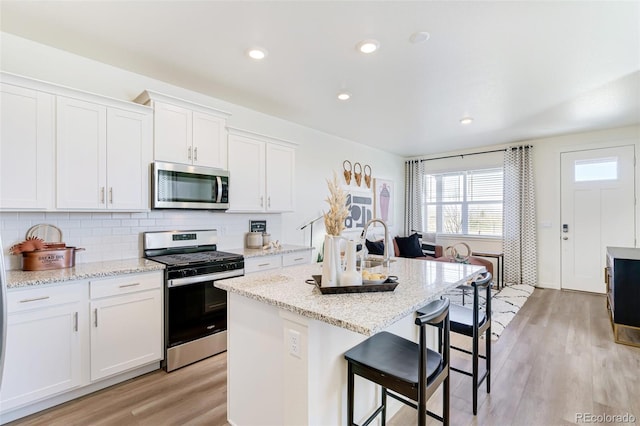 kitchen with light wood-type flooring, a kitchen island with sink, stainless steel appliances, white cabinets, and light stone counters