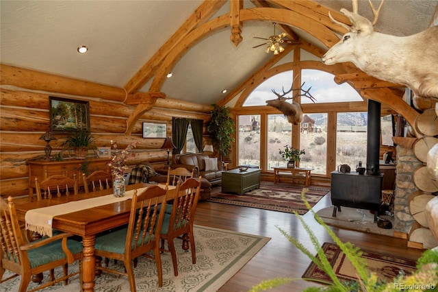 dining room featuring log walls, wood finished floors, beam ceiling, ceiling fan, and a wood stove