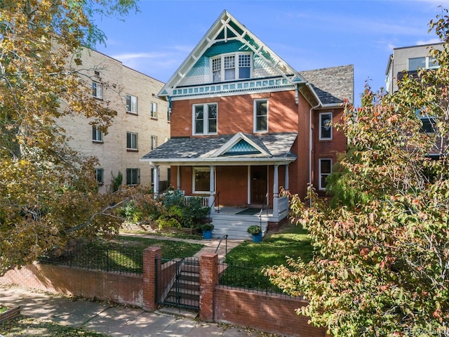 victorian-style house featuring a fenced front yard, roof with shingles, covered porch, a gate, and brick siding