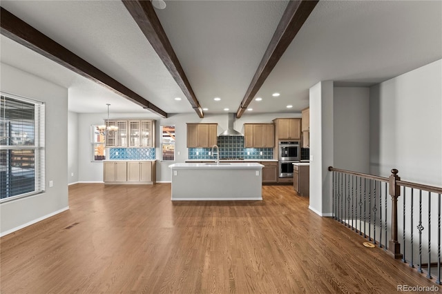 kitchen with decorative light fixtures, hardwood / wood-style floors, beamed ceiling, a kitchen island with sink, and wall chimney range hood