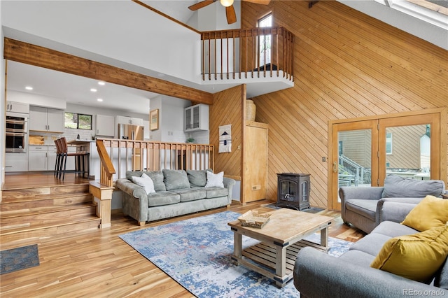 living area featuring a high ceiling, a wood stove, wood walls, a ceiling fan, and light wood-type flooring