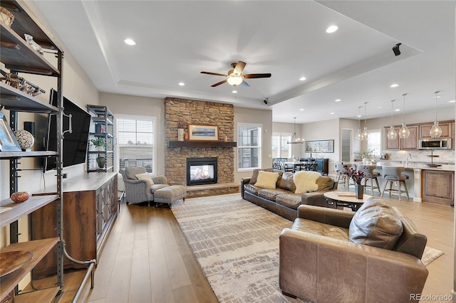 living room with ceiling fan with notable chandelier, a fireplace, a raised ceiling, and light wood-type flooring