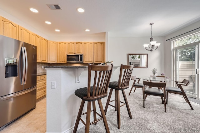 kitchen featuring light brown cabinetry, stainless steel appliances, an inviting chandelier, dark countertops, and a kitchen bar