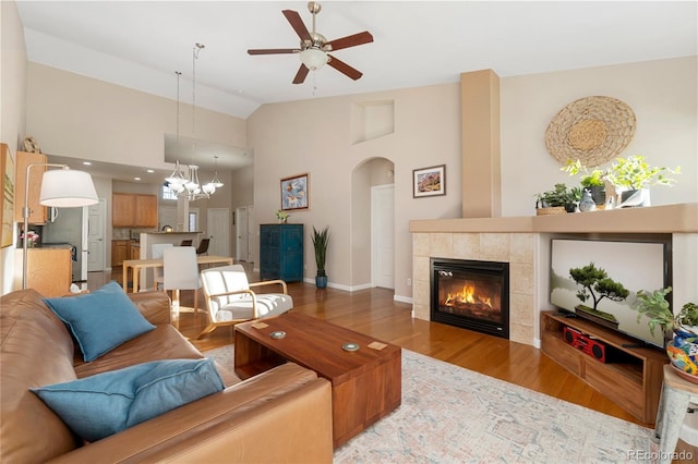 living room with arched walkways, a tiled fireplace, wood finished floors, high vaulted ceiling, and ceiling fan with notable chandelier