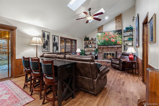 living area featuring vaulted ceiling with skylight, a brick fireplace, light wood-style flooring, and wainscoting