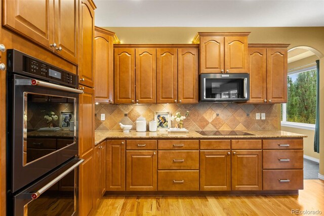 kitchen with black electric cooktop, stainless steel double oven, light stone countertops, light wood-type flooring, and brown cabinets