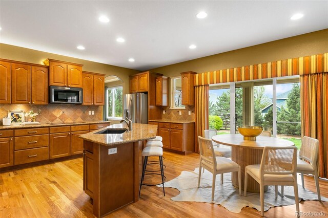 kitchen with appliances with stainless steel finishes, a sink, brown cabinets, light stone counters, and a kitchen island with sink