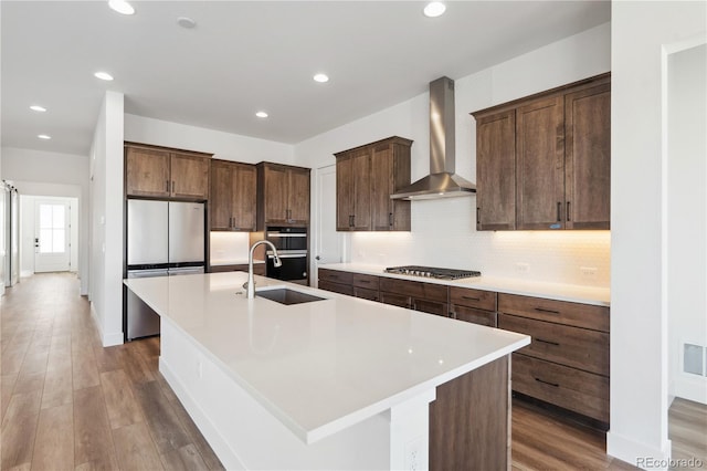 kitchen with wall chimney exhaust hood, a kitchen island with sink, stainless steel appliances, and light countertops