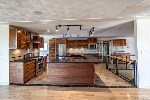 kitchen featuring light wood-type flooring, stainless steel appliances, a center island with sink, and wall chimney range hood