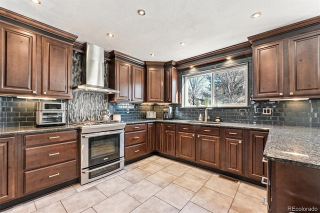 kitchen with tasteful backsplash, wall chimney exhaust hood, dark stone countertops, stainless steel range oven, and a sink