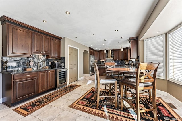 kitchen featuring arched walkways, wall chimney range hood, dark brown cabinets, beverage cooler, and stainless steel fridge with ice dispenser