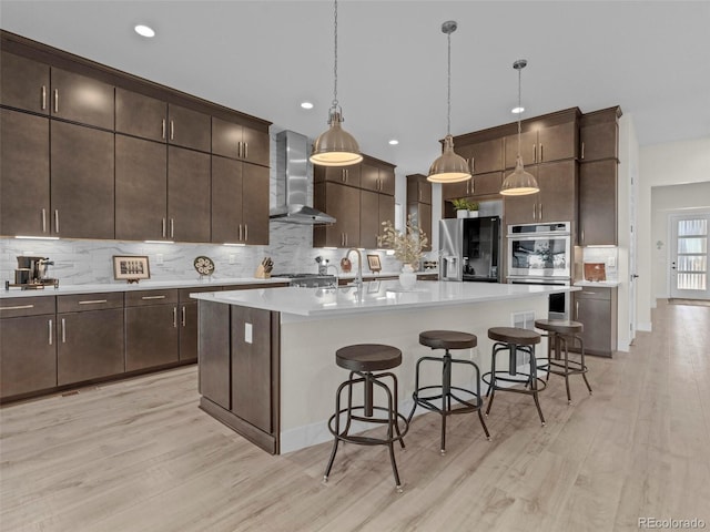 kitchen with a center island with sink, wall chimney range hood, hanging light fixtures, double oven, and refrigerator with ice dispenser