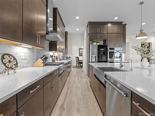 kitchen featuring wall chimney range hood, hanging light fixtures, light hardwood / wood-style floors, light stone counters, and stainless steel appliances