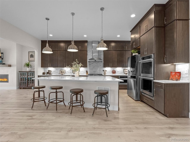 kitchen featuring dark brown cabinetry, stainless steel appliances, wall chimney range hood, tasteful backsplash, and decorative light fixtures