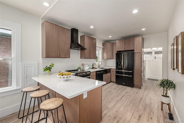 kitchen with a breakfast bar area, wall chimney range hood, light countertops, black appliances, and a peninsula