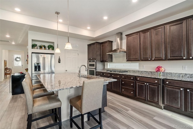 kitchen with wall chimney range hood, light stone countertops, a breakfast bar, stainless steel appliances, and a sink