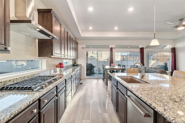 kitchen featuring a tray ceiling, a sink, light wood-style floors, appliances with stainless steel finishes, and wall chimney exhaust hood