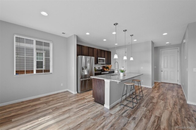 kitchen featuring light stone countertops, dark brown cabinets, stainless steel appliances, hanging light fixtures, and an island with sink