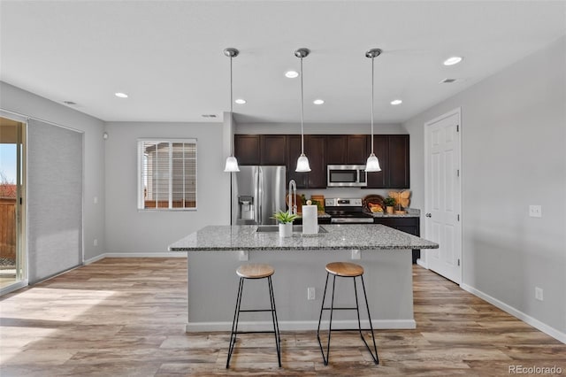 kitchen with light stone countertops, dark brown cabinetry, stainless steel appliances, a center island with sink, and a breakfast bar area