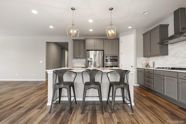 kitchen with dark wood finished floors, stainless steel appliances, gray cabinetry, a kitchen island with sink, and wall chimney range hood