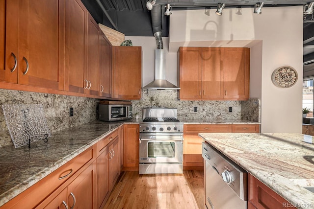 kitchen featuring wall chimney exhaust hood, appliances with stainless steel finishes, light stone countertops, and brown cabinets