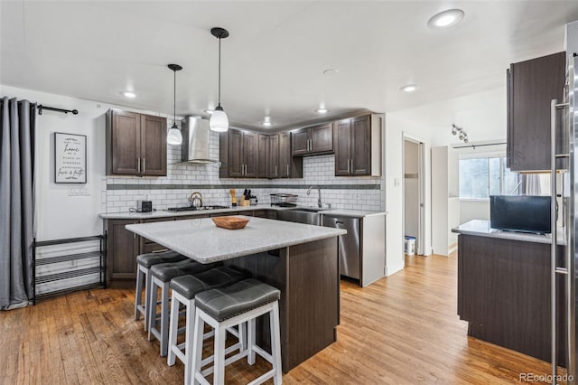 kitchen featuring light wood-type flooring, stainless steel appliances, wall chimney range hood, a kitchen island, and dark brown cabinetry