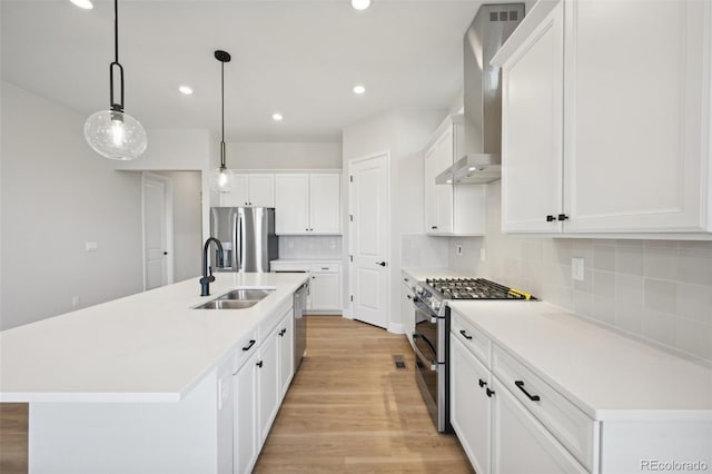 kitchen featuring hanging light fixtures, wall chimney range hood, light hardwood / wood-style flooring, a kitchen island with sink, and appliances with stainless steel finishes