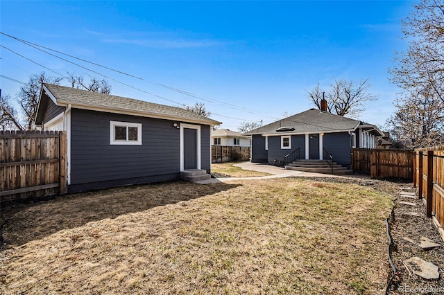 back of house with entry steps, roof with shingles, a lawn, a fenced backyard, and an outbuilding