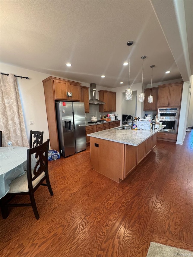 kitchen with appliances with stainless steel finishes, dark hardwood / wood-style floors, decorative light fixtures, a kitchen island with sink, and wall chimney exhaust hood