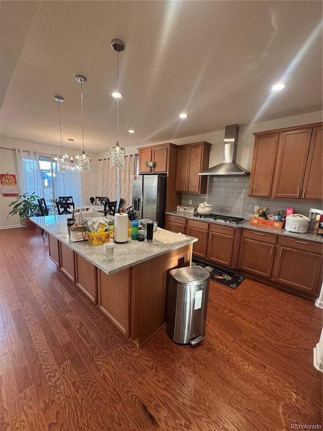 kitchen with pendant lighting, wall chimney range hood, dark wood-type flooring, a kitchen island with sink, and stainless steel appliances