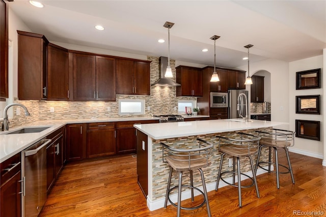 kitchen with sink, a center island with sink, wall chimney range hood, and appliances with stainless steel finishes