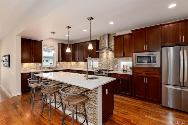 kitchen featuring stainless steel appliances, a kitchen island with sink, wall chimney range hood, decorative light fixtures, and dark hardwood / wood-style floors