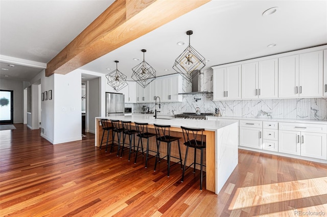 kitchen featuring stainless steel refrigerator, a kitchen island with sink, white cabinets, and wall chimney range hood