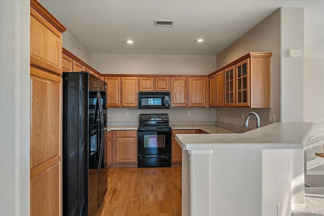 kitchen with light countertops, glass insert cabinets, black appliances, a peninsula, and light wood-style flooring