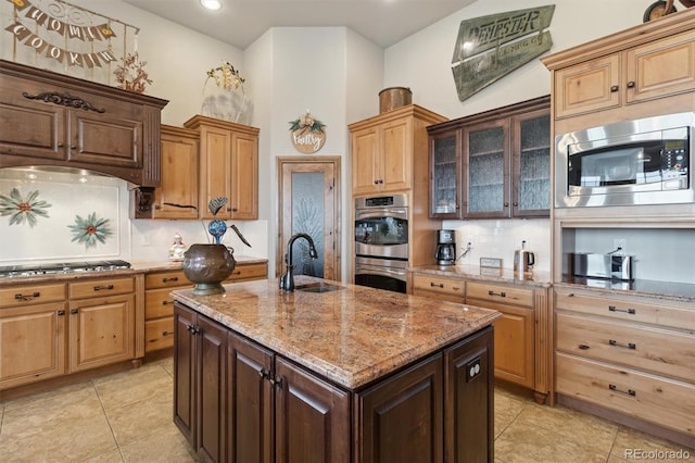 kitchen with appliances with stainless steel finishes, sink, backsplash, light stone countertops, and a center island with sink