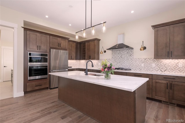 kitchen featuring wall chimney range hood, sink, dark brown cabinets, stainless steel appliances, and light stone countertops