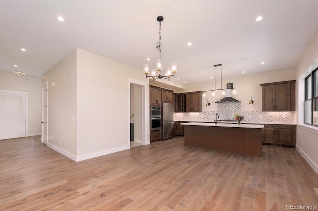 kitchen with wall chimney range hood, appliances with stainless steel finishes, a kitchen island with sink, backsplash, and decorative light fixtures