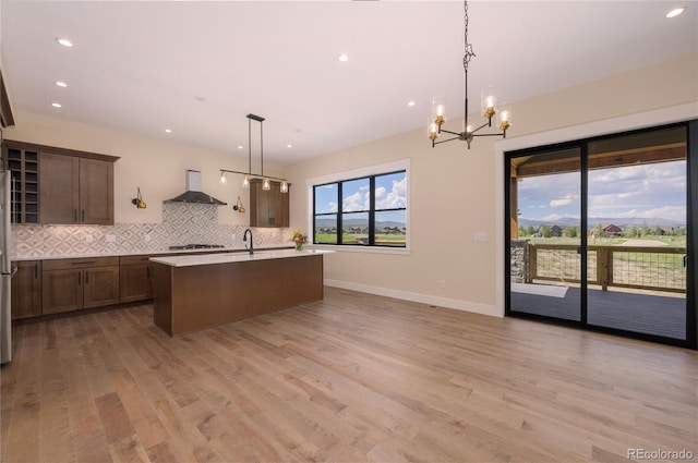 kitchen with wall chimney range hood, light hardwood / wood-style flooring, hanging light fixtures, tasteful backsplash, and a center island with sink