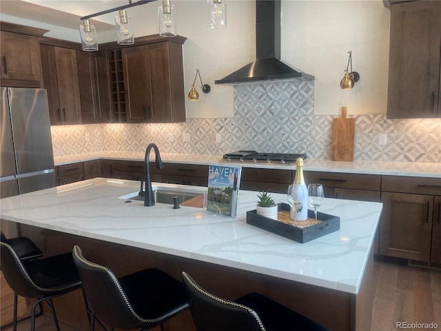 kitchen featuring wall chimney range hood, sink, stainless steel appliances, light stone counters, and decorative backsplash