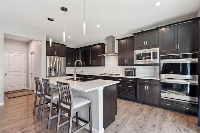 kitchen featuring appliances with stainless steel finishes, a kitchen island with sink, sink, wall chimney range hood, and decorative light fixtures